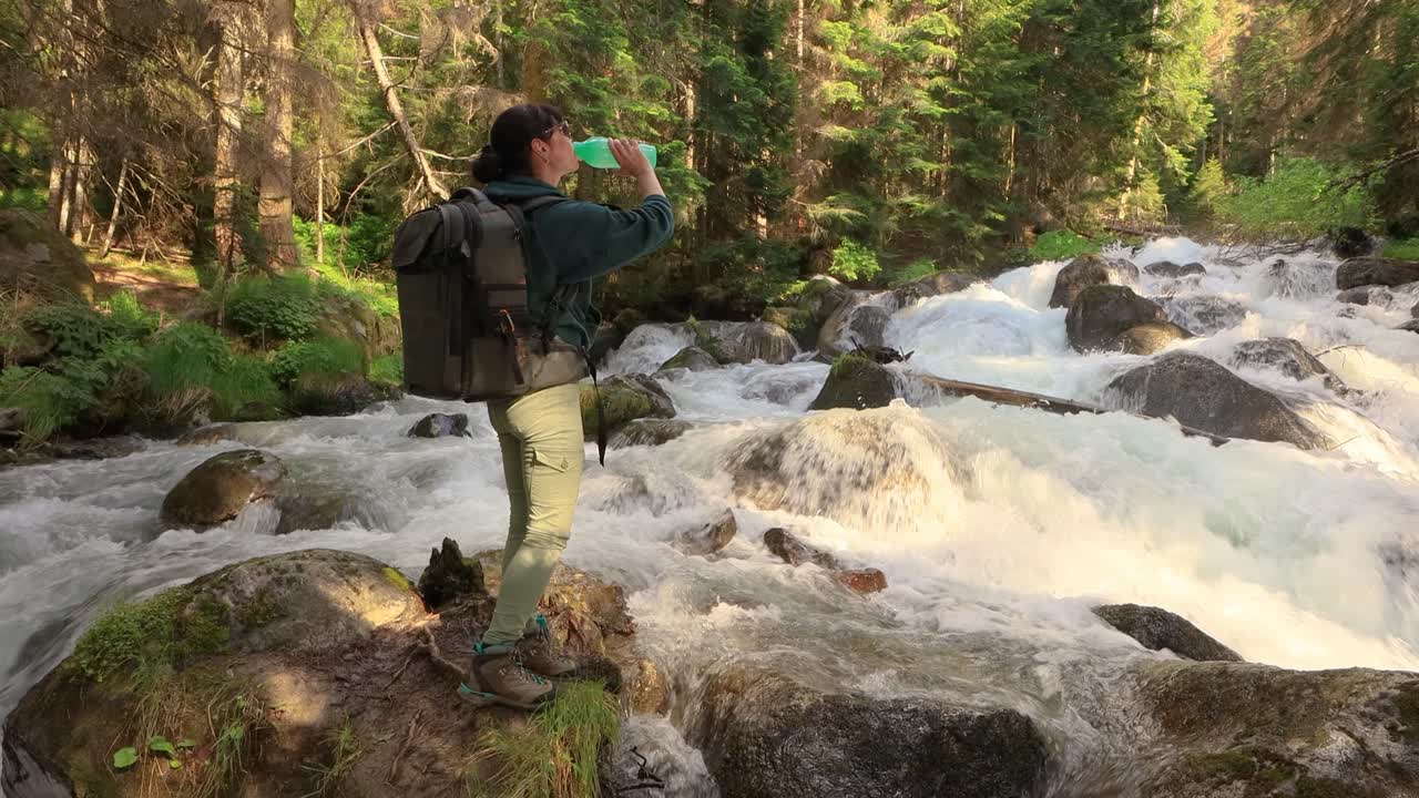 viajera con una mochila, bebiendo agua en la naturaleza en el bosque cerca de un río de montaña.
