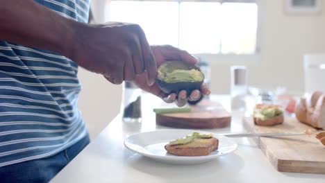 Hombre-Afroamericano-Poniendo-Aguacate-Sobre-Tostadas-En-Una-Cocina-Soleada,-Cámara-Lenta