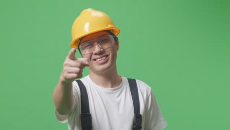 close up of asian man worker wearing goggles and safety helmet smiling and touching his chest then pointing at you while standing in the green screen background studio