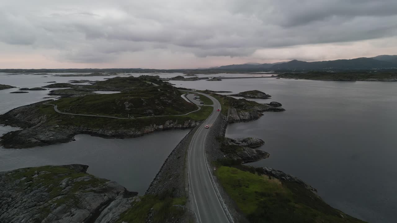 다양한 섬을 따라 이동하면서 먼 산을 덮고 있는 구름과 함께 여름 일몰에 노르웨이의 대서양 도로를 지나가는 자동차 위를 날고 있습니다.