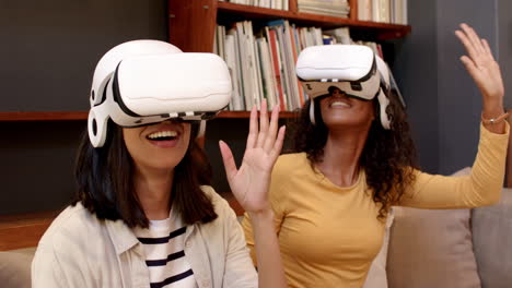 two multiracial female friends enjoying virtual reality experience at home, smiling and waving hands