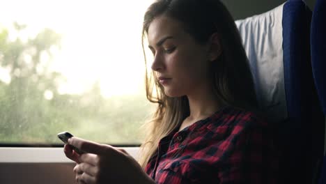 Attractive-female-in-plaid-shirt-is-going-by-train,-sitting-next-to-the-window-and-seriously-looking-on-her-mobile-phone.-Side-view.-Transport,-motion