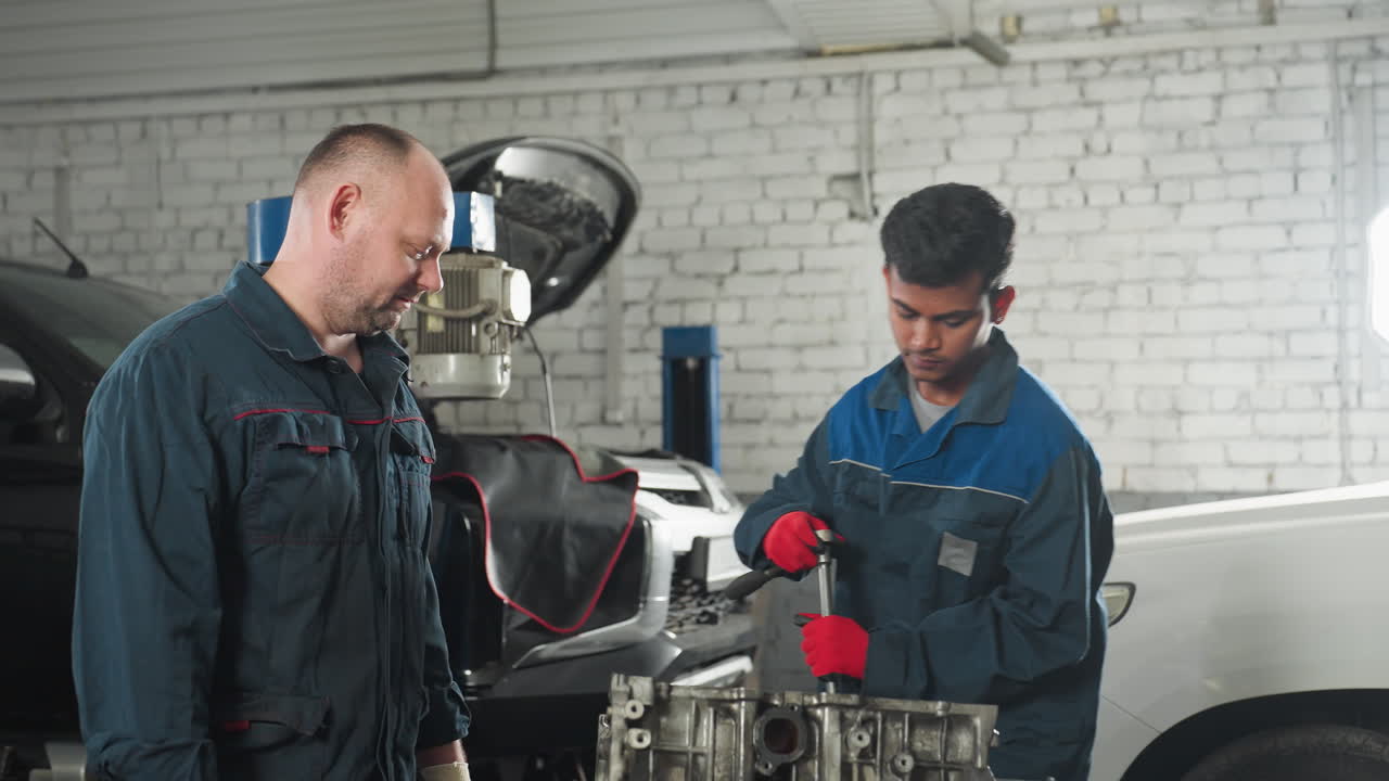 ingenieros automotores en un taller automotriz, uno con guantes rojos trabajando en un motor de coche usando una herramienta mientras un colega observa con la mano en el motor, el fondo presenta vehículos