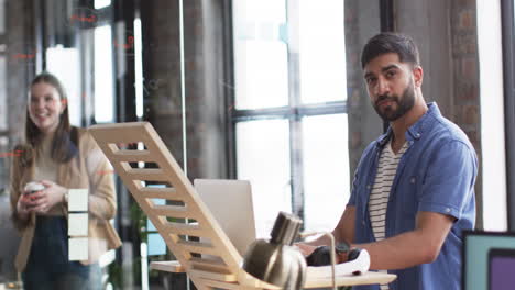 Young-Caucasian-woman-smiles-in-the-background-as-young-Asian-man-focuses-on-his-work-in-a-business-