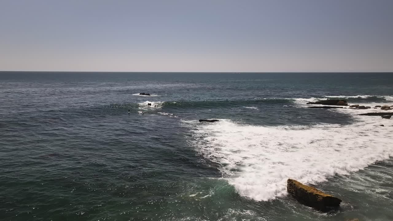 surfistas en la playa de la laguna tratando de atrapar una ola en el océano pacífico con algunas rocas, cielo azul y sin nubes