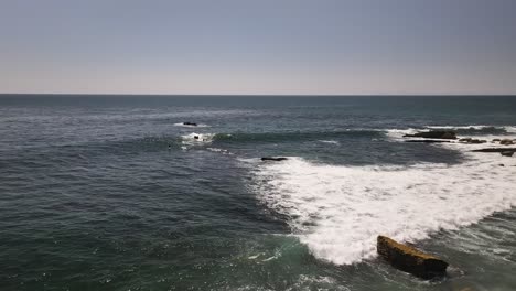 Surfers-in-Laguna-Beach-trying-to-catch-a-wave-in-the-Pacific-Ocean-with-some-rocks,-blue-sky-and-no-clouds