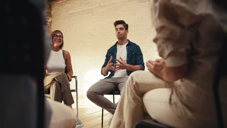 a brunette man in a white t-shirt and denim jacket expresses his thoughts in group therapy. other group therapy participants