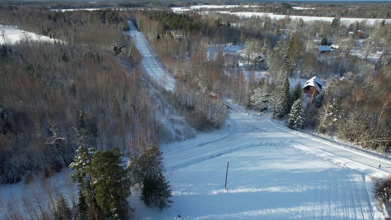 Aerial view of a serene winter countryside featuring snow-covered trees, wooden houses, and a winding road through a frosty forest.