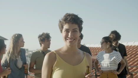 retrato de una mujer caucásica feliz con el pelo corto sosteniendo una cerveza y mirando a la cámara mientras tiene una fiesta en la azotea con amigos