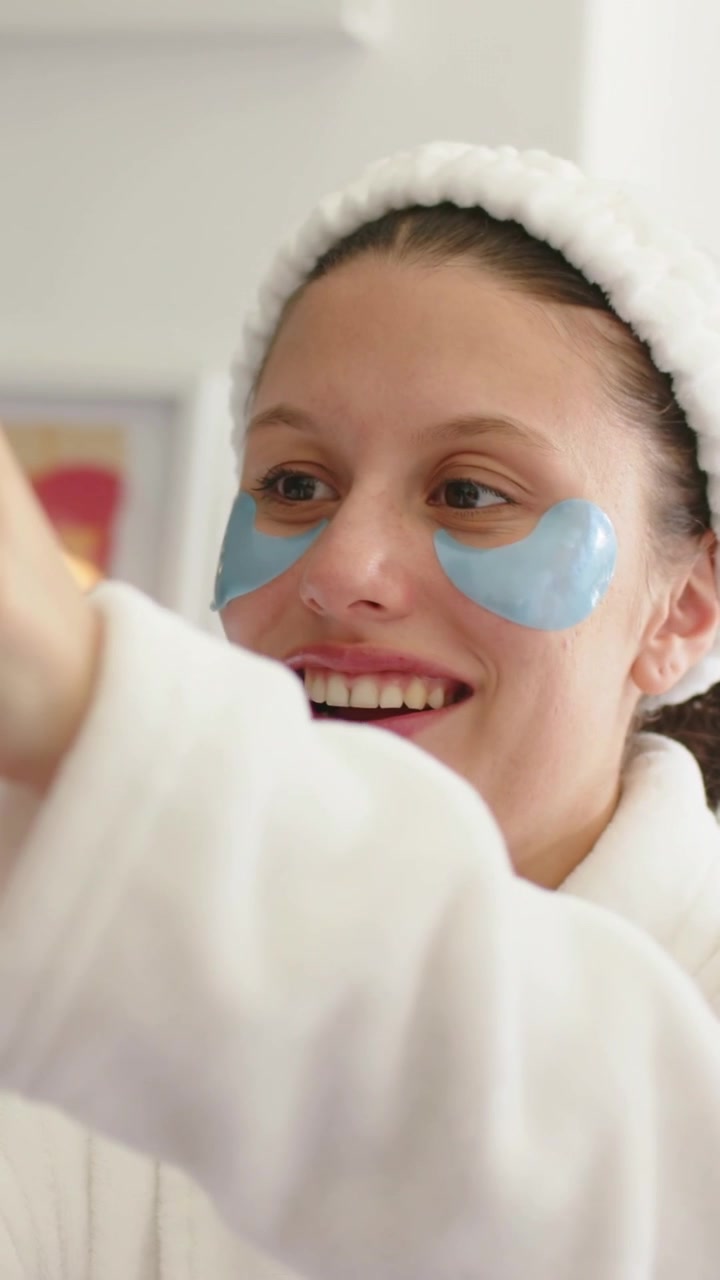 Vertical video: Cam focusing, woman reaching right hand at vanity, robe, headband, blue patches