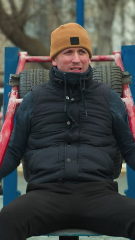 a coach seated on workout equipment outdoors, resting his hands on red rods, looks tired as he looks to the left, in the background, a boy dressed in black plays football