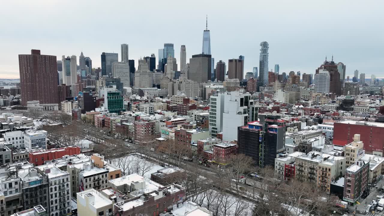 Bredt luftfoto af New York City om vinteren, der viser Manhattans skyline med tætte skyskrabere, sneklædte tage, boligbyggerier og nøgne træer under en overskyet himmel. Vinterscene