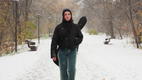 lady in winter jacket walking during snow, another person following, snowy park path lined with lamp posts and benches, tranquil winter scene with falling snowflakes, trees retaining autumn leaves