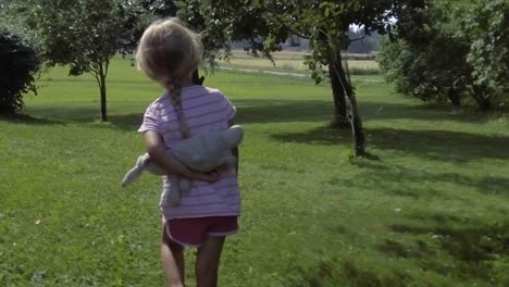 three year old summer girl walking on grass field among apple trees, holding stuffed animal, pink clothes, slow motion