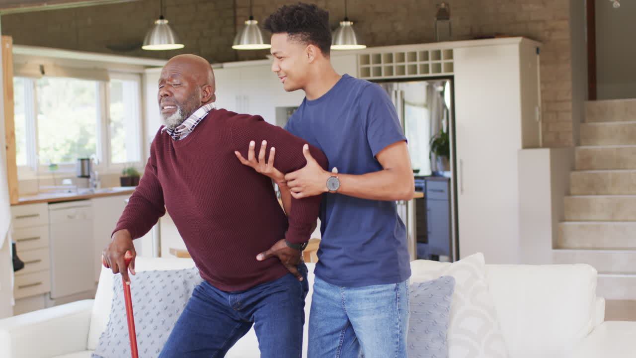 Happy african american adult son helping senior father stand up from couch at home, slow motion