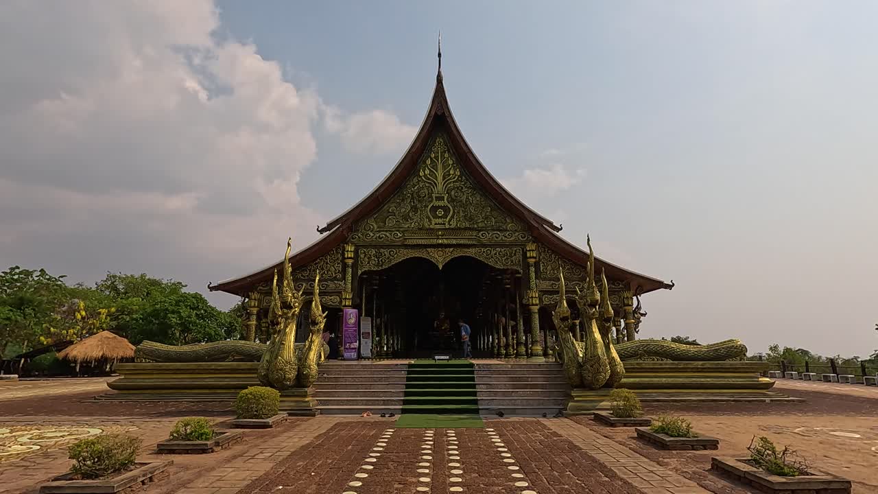 Camera slowly pans toward a traditional Buddhist temple with intricate architecture, green landscaping, and a patterned walkway under soft daylight and partly cloudy skies