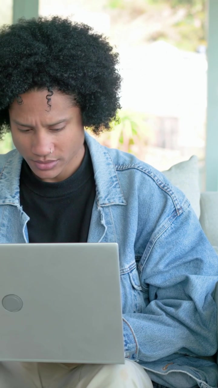 Mid adult male wearing denim jacket carrying silver laptop sitting on covered porch sofa typing