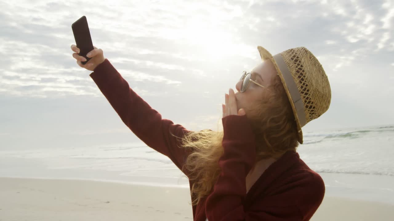 vista lateral de una joven y hermosa mujer caucásica haciendo clic en una selfie con un teléfono móvil en la playa 4k