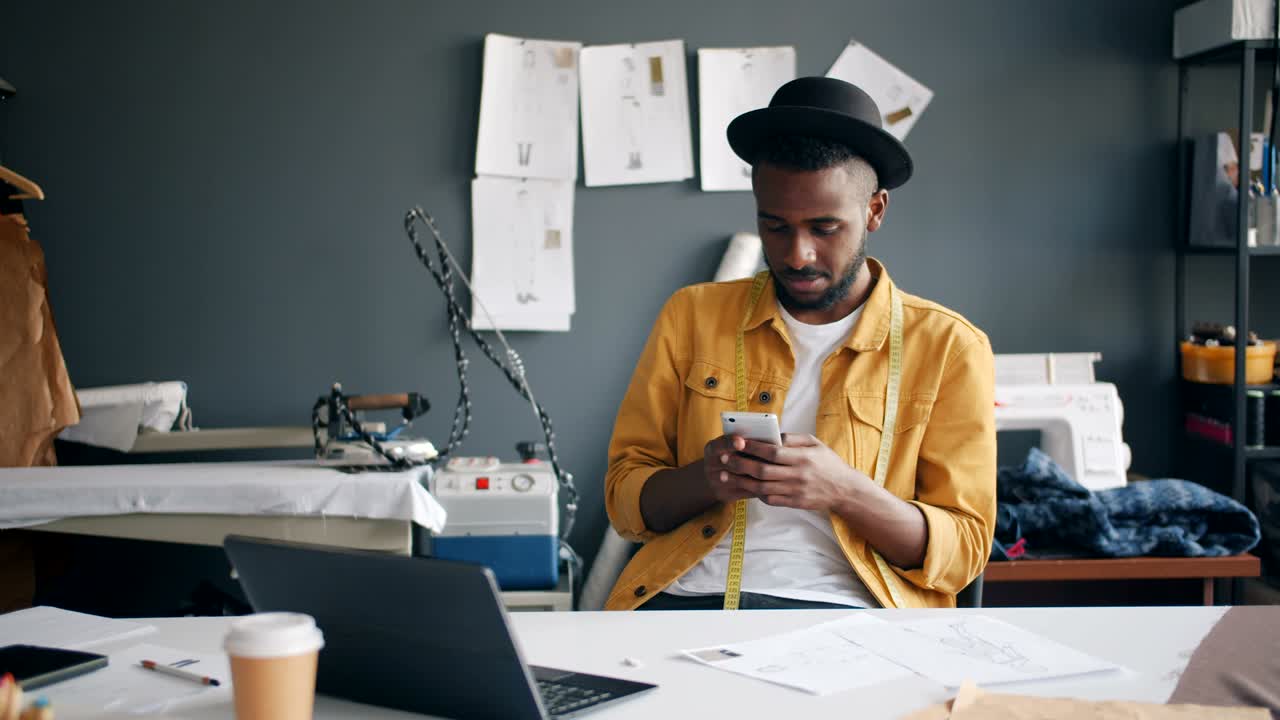 African American fashion designer using smartphone smiling at work in studio