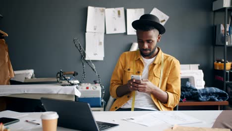 african american fashion designer using smartphone smiling at work in studio