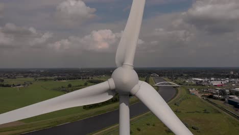 steady aerial view of a single wind turbine with rotating blades generating electricity and dutch waterway landscape in the background