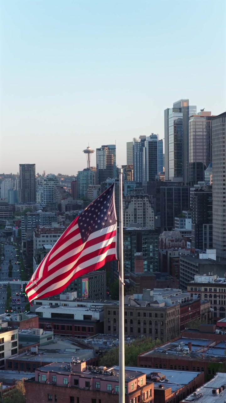 Bandiera americana sventola davanti allo skyline con lo Space Needle a Seattle, Washington, dove dense torri condominiali in vetro si raggruppano strettamente sotto la luce dorata e velata del tramonto serale. Ripresa aerea verticale con drone.