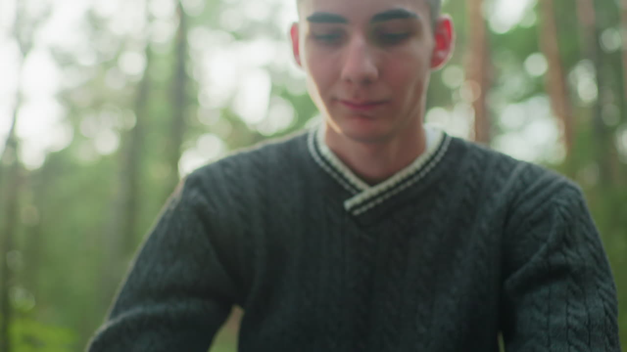 close up of young boy with serious expression looking downward in forest wearing dark knitted sweater and light jeans as soft sunlight filters through trees