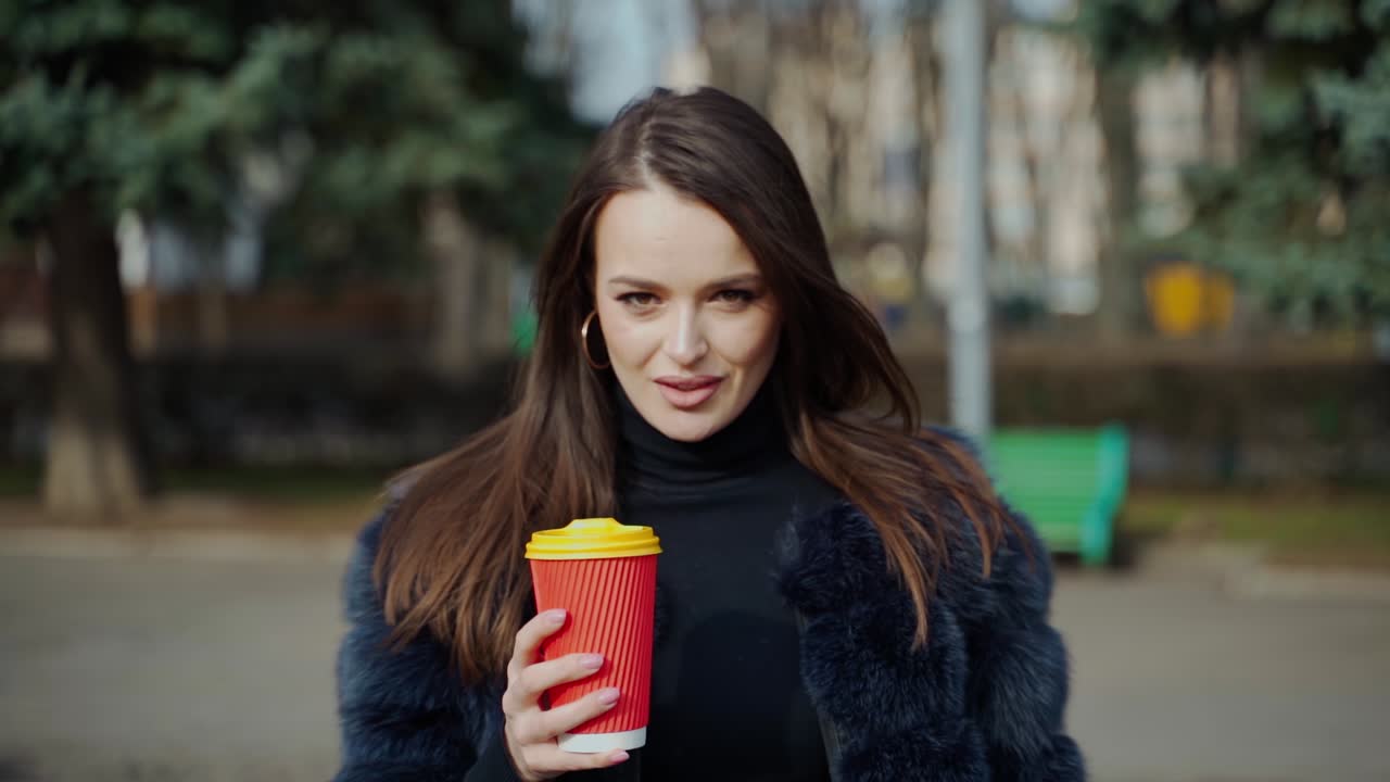 Portrait of seductive woman. Beautiful girl in fluffy coat holds plastic cup of hot drink and looks on camera in the city park.