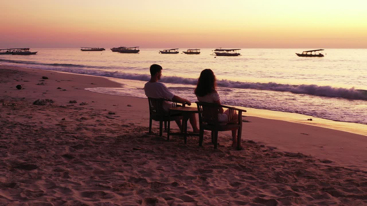 Young couple relaxing on calm exotic beach, sitting on chairs over white sand, watching beautiful sunset with warm sunlight