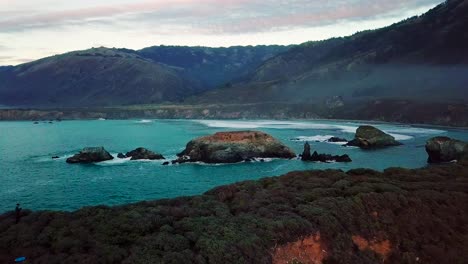 Vista-Aérea-Crepuscular-De-Enormes-Acantilados-Oceánicos-Y-Olas-Rompiendo-En-Las-Rocas-En-La-Playa-Del-Dólar-De-Arena-En-Big-Sur-California