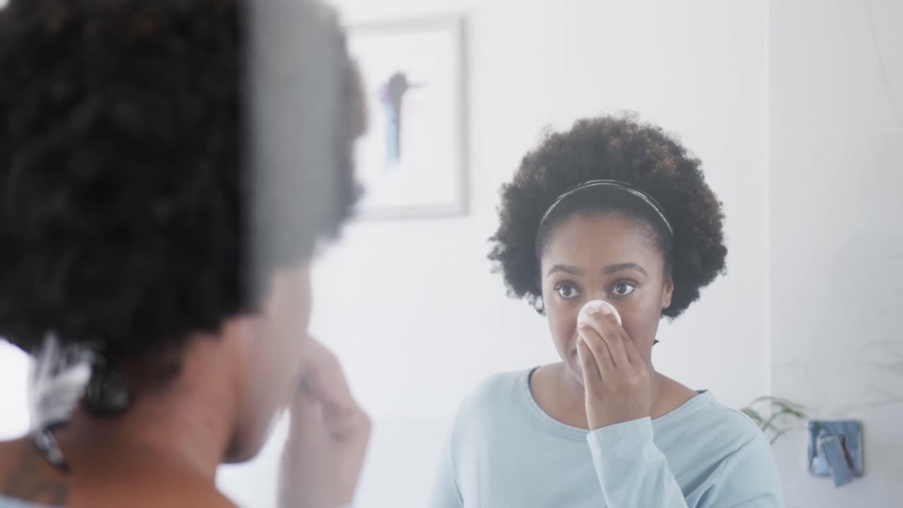 feliz mujer afroamericana limpiando la cara con una almohadilla de algodón, mirando en el espejo del baño, en cámara lenta