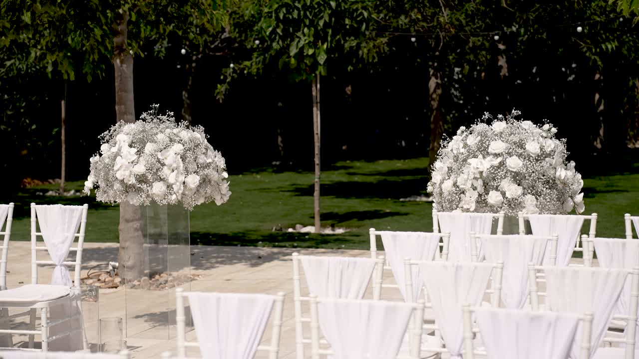 A charming garden is prepared for a wedding ceremony. Rows of white chairs face stunning floral arrangements. The setting is peaceful and provides a lovely backdrop for the event
