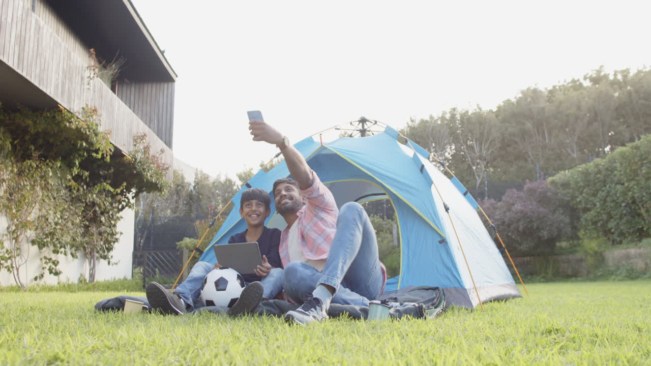 Taking selfie, Indian father and son sitting by tent with soccer ball and tablet