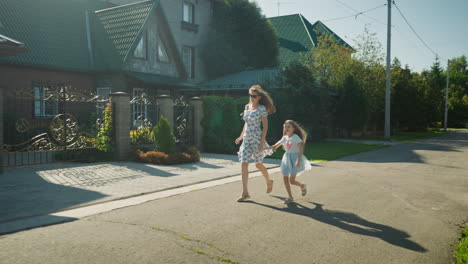 young woman in floral dress running with little girl in light dress along quiet sunny street, both appearing hurried as if late for event, passing green hedges and houses in peaceful neighborhood