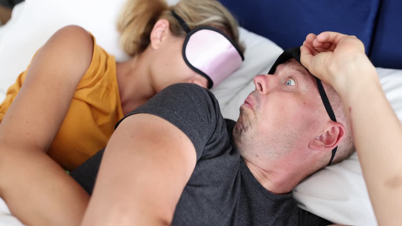 Man awake and worried in bed next to sleeping partner