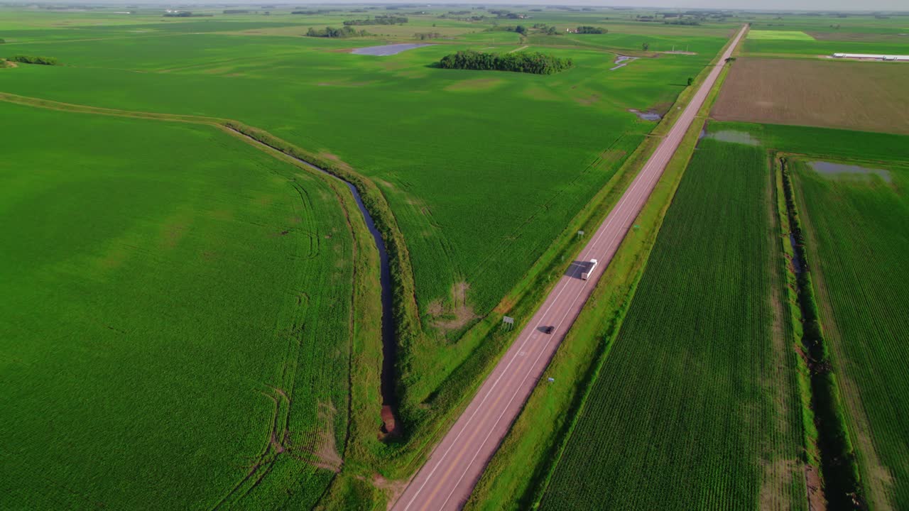 Aerial shot of semi-trucks with grains trailers traveling on a rural road through green farmland in Minnesota, highlighting transportation.