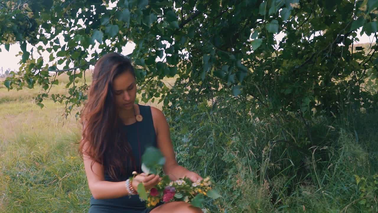 una hermosa joven vestida de negro y arreglando flores y hojas alrededor del árbol al aire libre durante la visita a los campos
