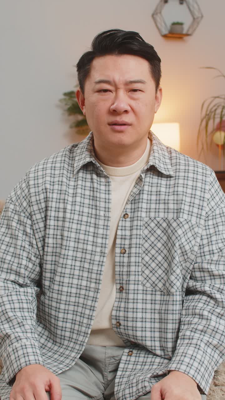 Confused asian handsome man feeling embarrassed about ambiguous question sitting on sofa at home