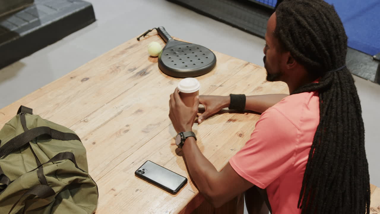 Man with smartphone and coffee sitting at table with padel racket and ball, on indoor court