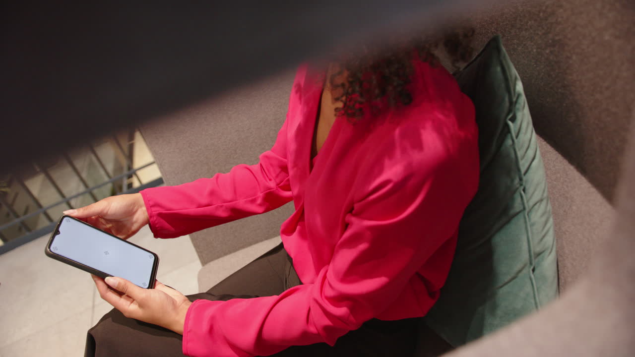 Holding smartphone, woman in pink blouse sitting on couch, working remotely, copy space
