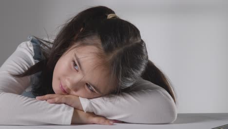 Studio-Shot-Of-Bored-Hyperactive-Girl-Sitting-At-Table-Against-White-Background