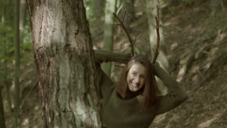 young woman peeks out from behind a tree, puts two sticks on her head and pretends to be a deer