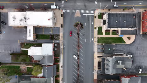 Aerial-tracking-of-red-car-driving-through-American-city,-past-USA-flag-in-town-square