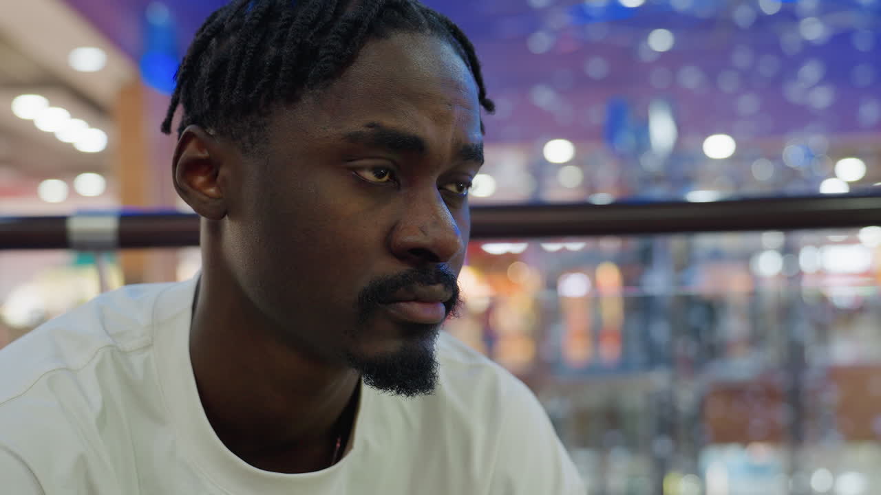 Close up of man seated in mall wearing white shirt with serious contemplative expression as he glances down thoughtfully and checks his time against blurred background lights