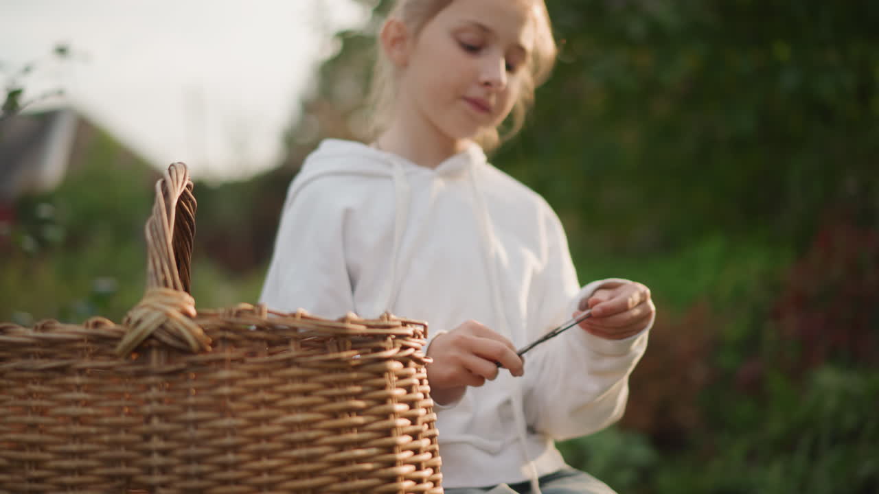 Bambina bianca all'aperto con un cesto di vimini, che ispeziona piccole forbici mentre è seduta su una panchina da giardino nella morbida luce dorata del pomeriggio, con indosso una felpa con cappuccio, espressione calma, mani concentrate che preparano un semplice lavoretto