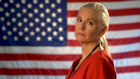 medium tight portrait of the back of blonde nurses head as she turns and looks at camera looking concerned and sad with out of focus us flag