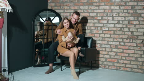 caucasian parents with little infant boy sit in armchair. portrait of a loving young family near brick wall.