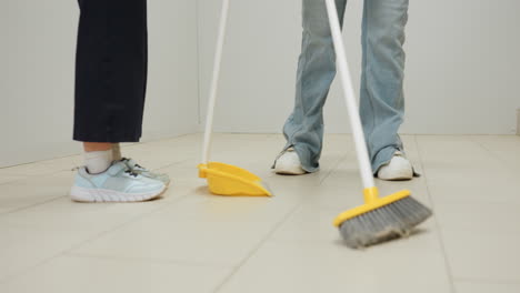 Close zoom of mother and child in laundromat, parent sweeping tiled floor with broom while kid holds dustpan, teamwork cleaning near industrial washers, family chore, hygiene focus, bonding