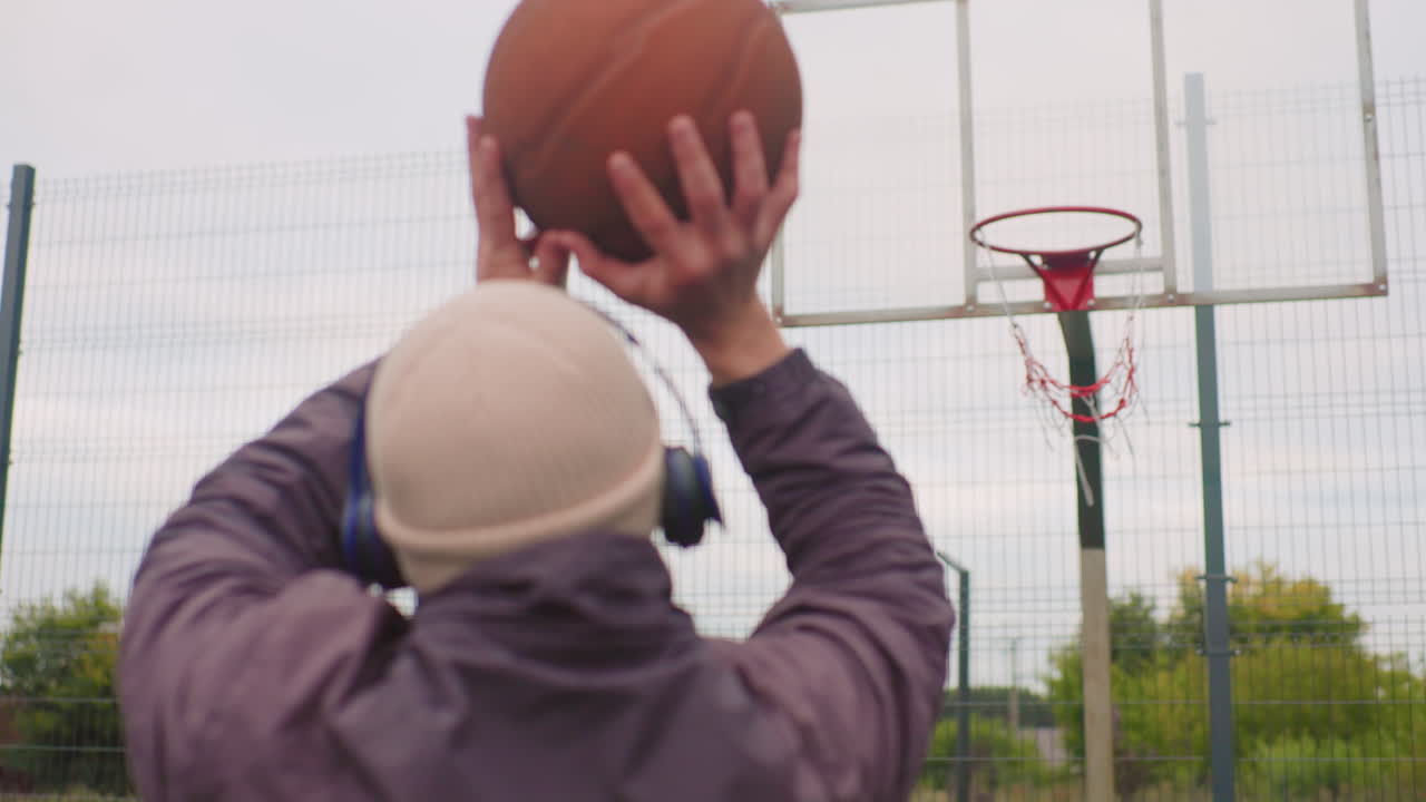 Persona che gioca a basket all'aperto con cuffie, berretto e giacca, che mira al canestro su un campo recintato sotto un cielo nuvoloso; il rilascio al rallentatore mostra un arco alto verso il bordo e la rete, allenamento concentrato