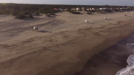 Disparo-Aéreo-En-Cámara-Lenta-De-Dos-Personas-Caminando-Sobre-La-Arena-Frente-A-Las-Olas-De-Agua-De-La-Playa-En-Un-Día-Nublado-Al-Atardecer-En-Mar-De-Las-Pampas,-Argentina